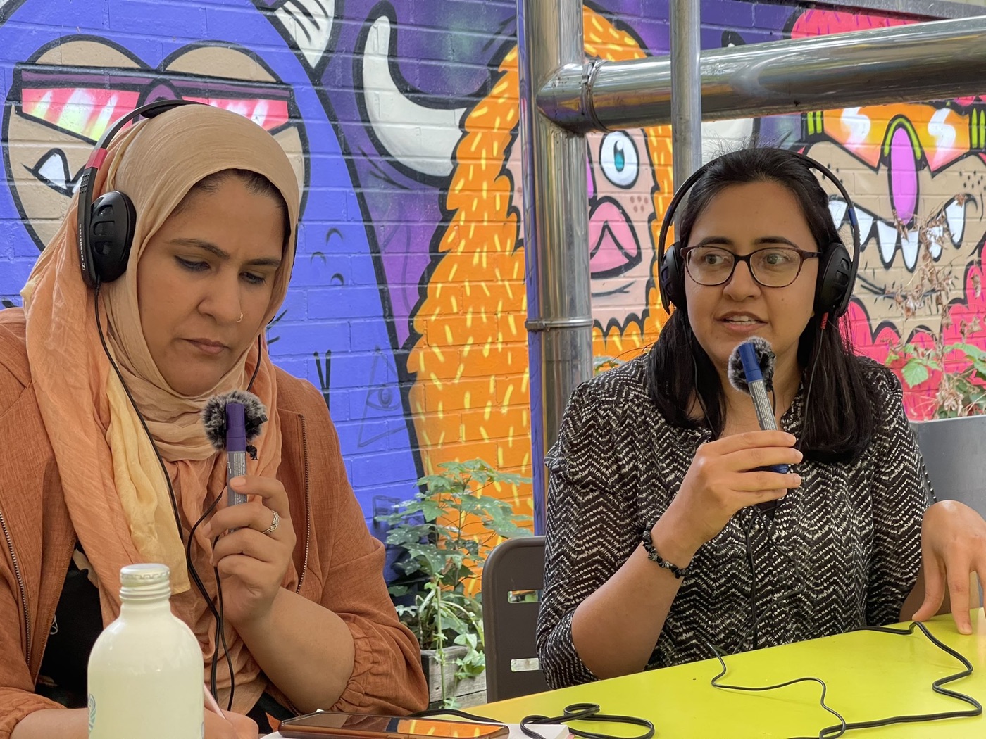 Two community reporters wearing headphones and holding microphones, recording a podcast outdoors in front of a brightly coloured graffiti mural.
