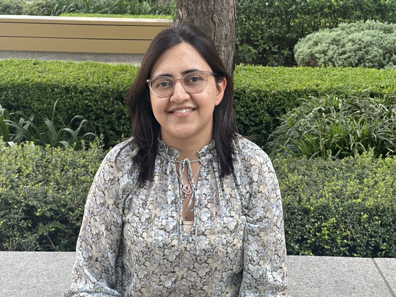 Dr Swati Virmani seated outdoors in front of a clipped hedge and a tree, smiling towards the camera.